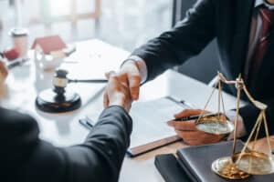 Lawyer shaking hands with a client across a desk with legal documents and scales of justice.