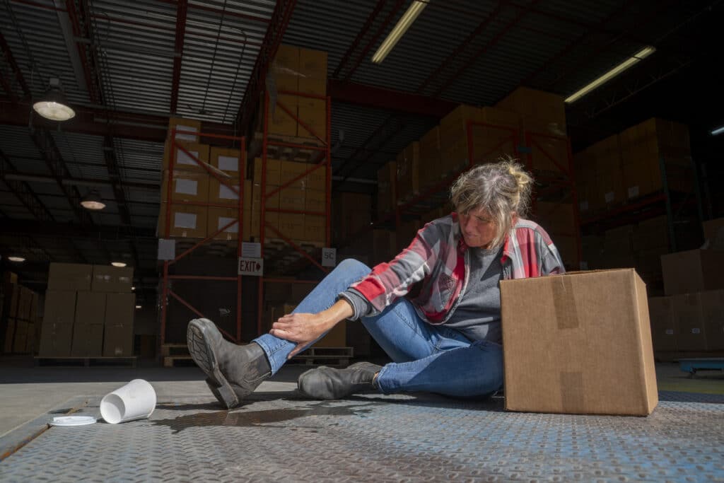 Warehouse worker holding an injured ankle after slipping near a spilled drink and cardboard box.